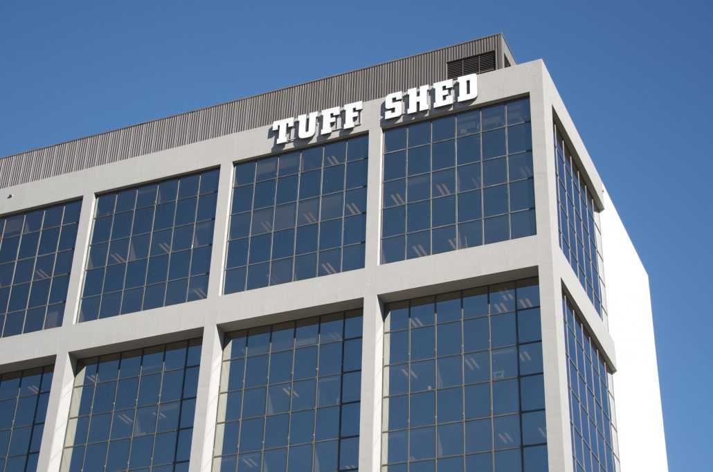 Exterior shot of the top of Tuff Shed headquarters taken from the parking garage aimed up focused on the large letters of the Tuff Shed sign and top 12 floors of the building and penthouse against a blue sky.