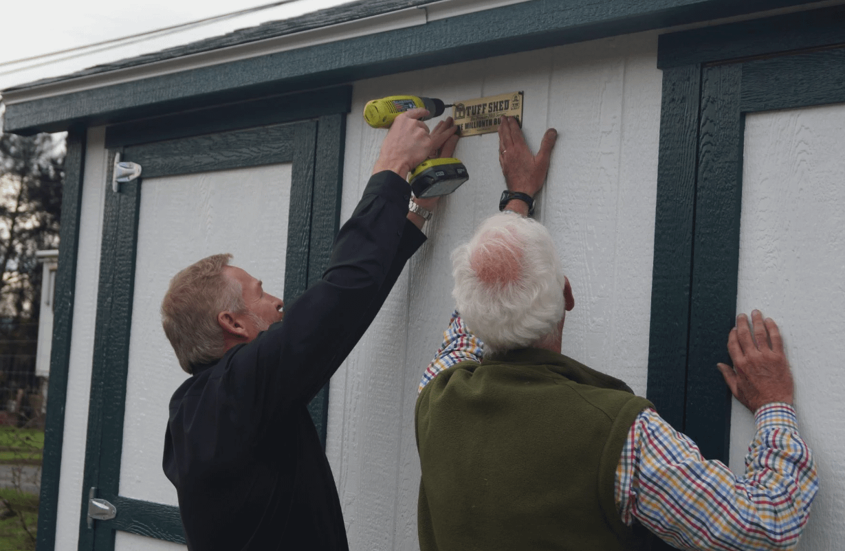 Exterior of a Tuff Shed Premier PRO Ranch white in color with two men installing the gold plated serial tag with a green drill onto the wall between two doors