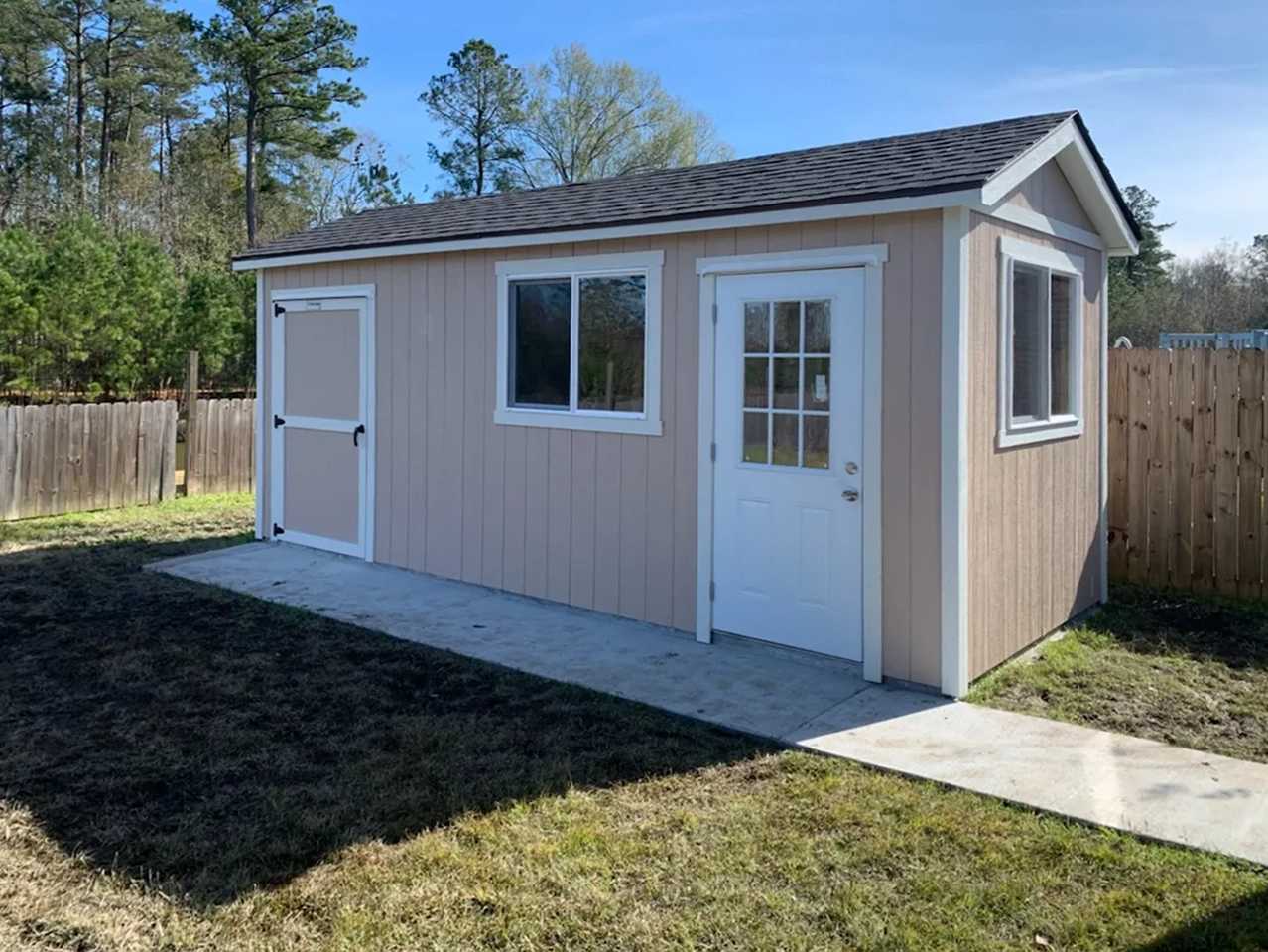 A small beige shed with a white door and windows stands on a concrete slab, surrounded by a wooden fence and trees under a clear blue sky.