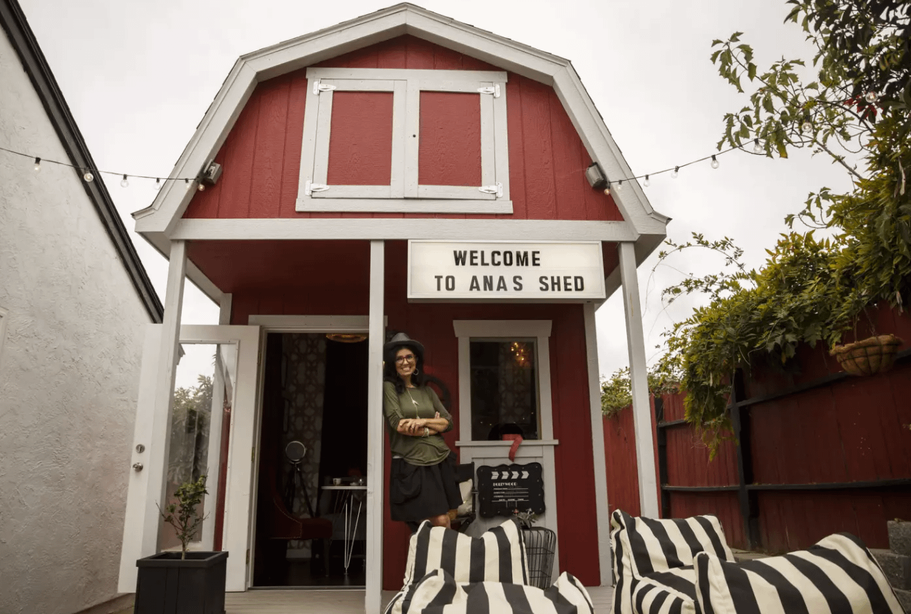Exterior of PRO Barn Weekender red in color with woman standing on porch leaning against pole there is a sign that says welcome to ana's shed