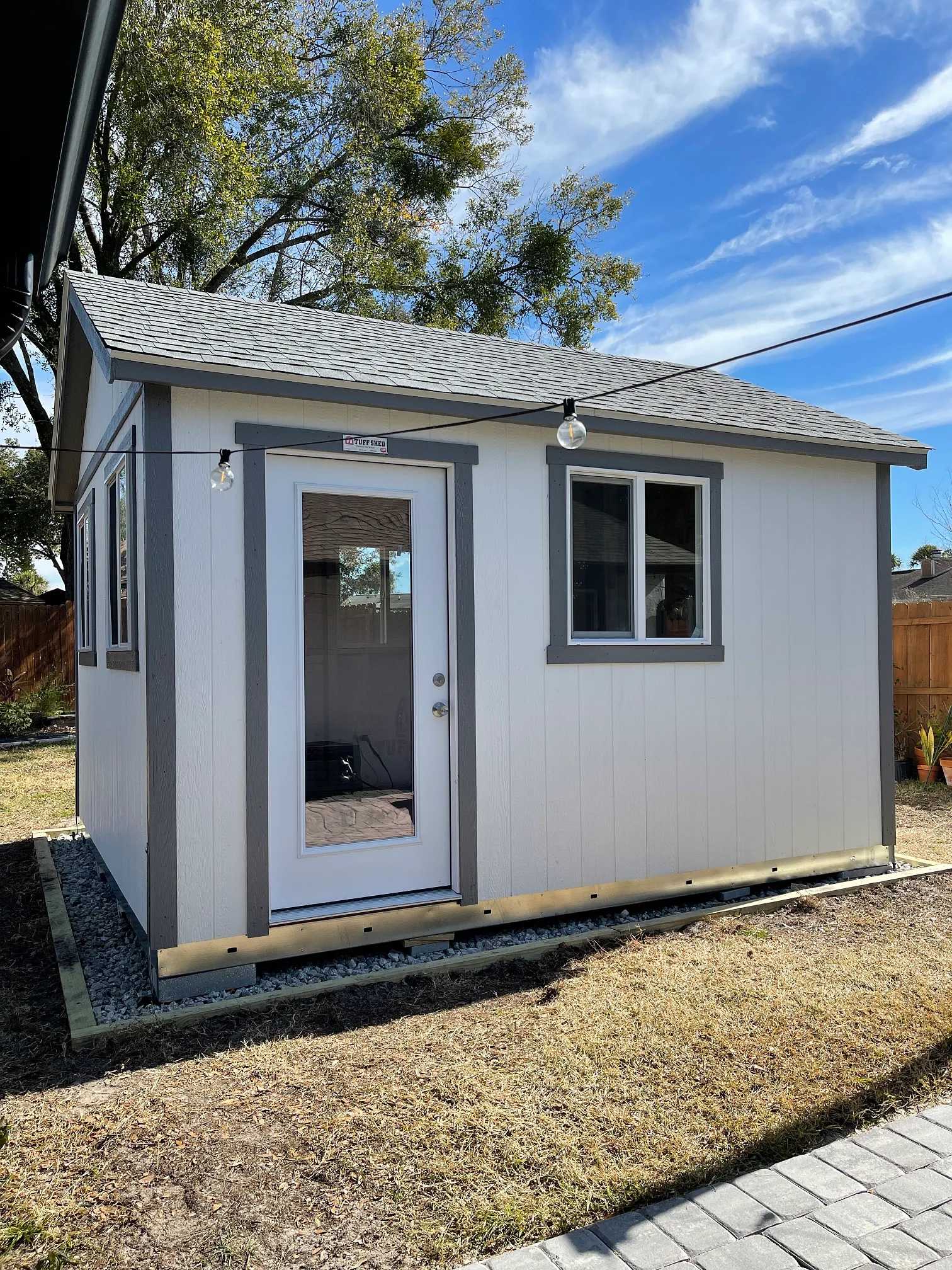 A small gray and white shed with a glass door and window sits in a yard under a blue sky with scattered clouds.