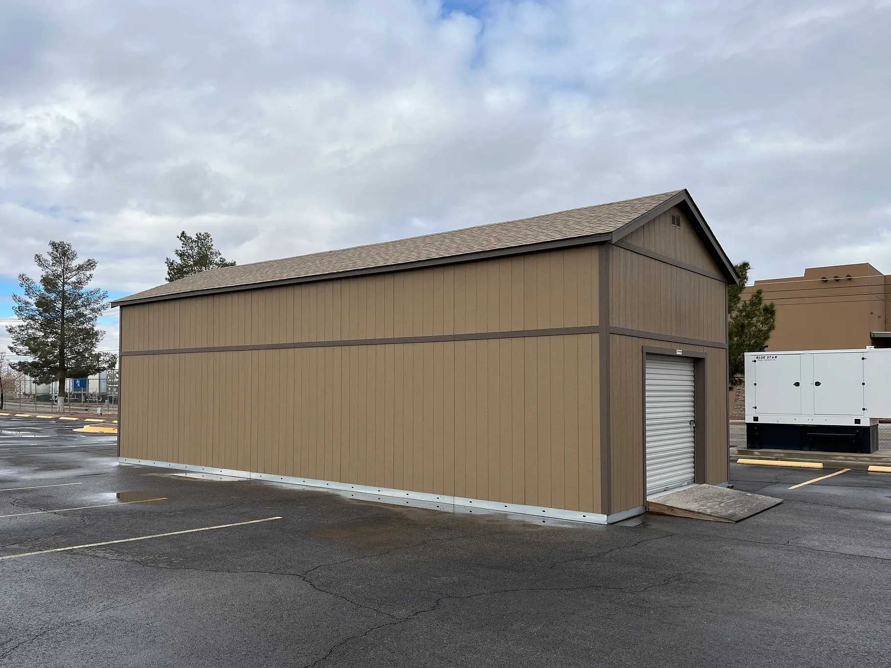 A tan storage shed with a roll-up door stands on a wet asphalt surface, surrounded by a cloudy sky and some trees in the background.
