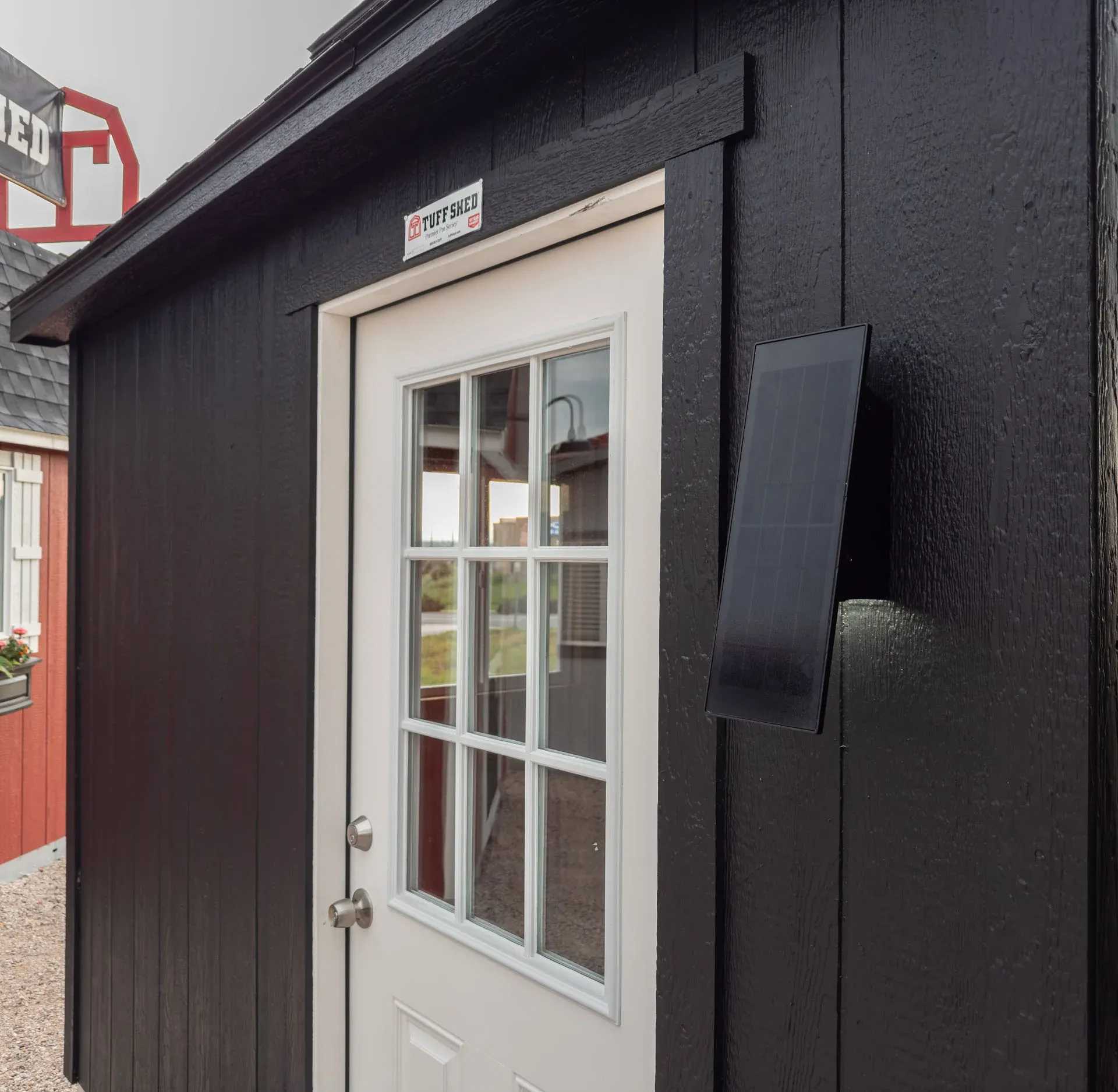 Black shed with a white door featuring glass panels, a solar panel attached to the wall, and a logo above the door.