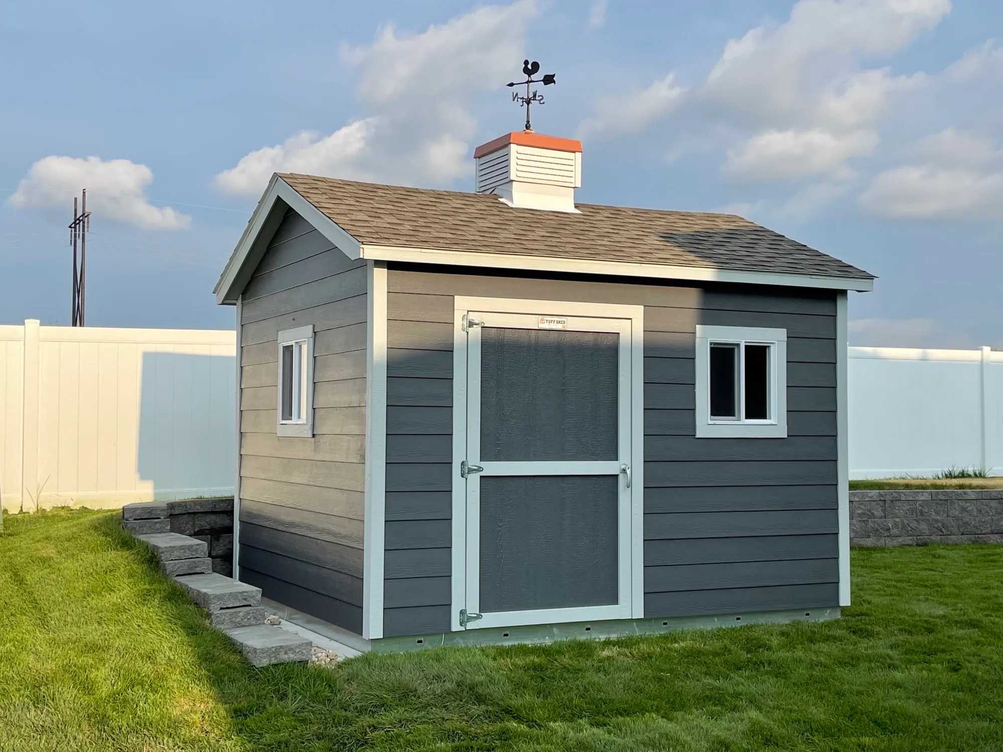 A small gray garden shed with windows, a white door, and a weather vane on top, set on lush green grass with a white fence in the background.
