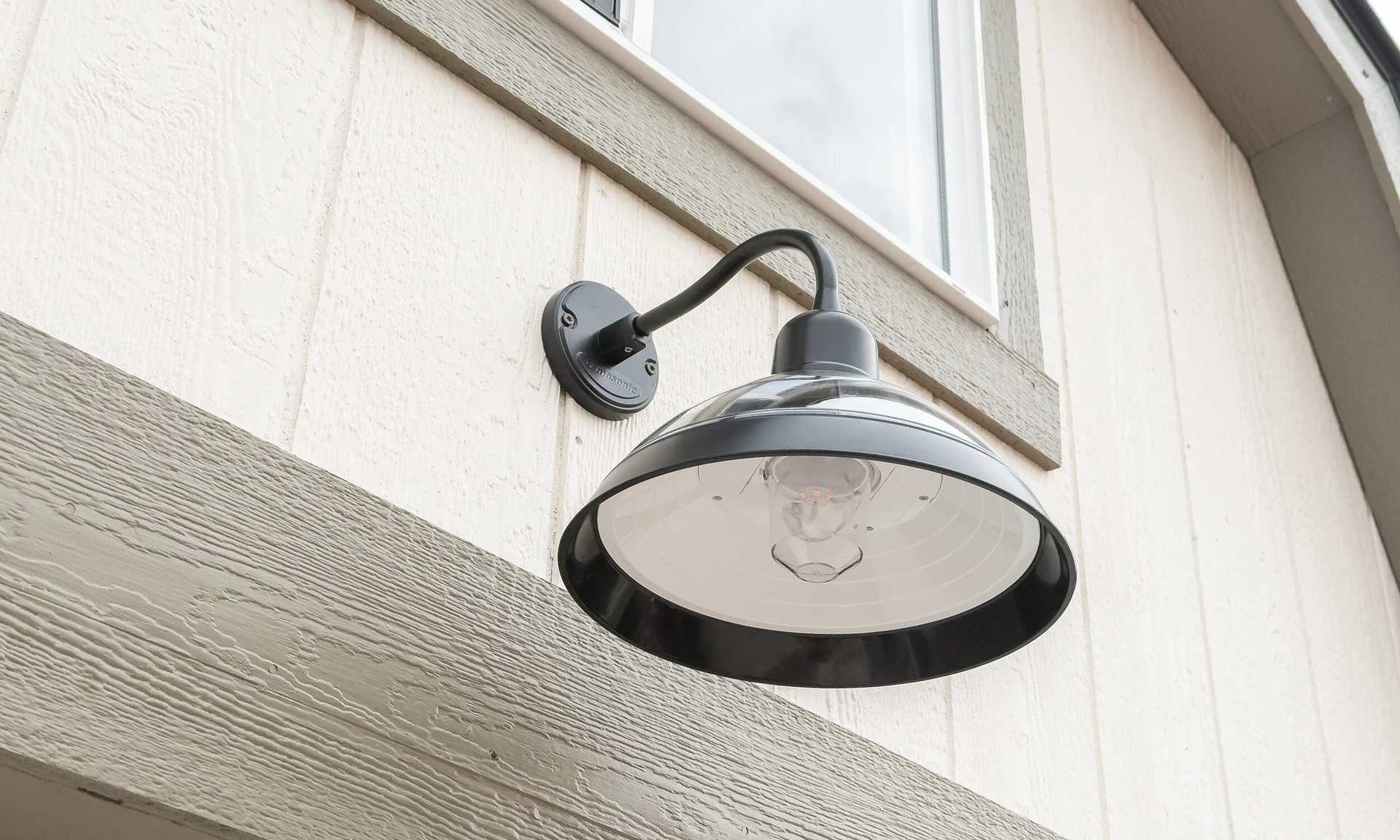 A black outdoor wall light fixture with a wide shade, mounted on a light-colored textured wall near a window.