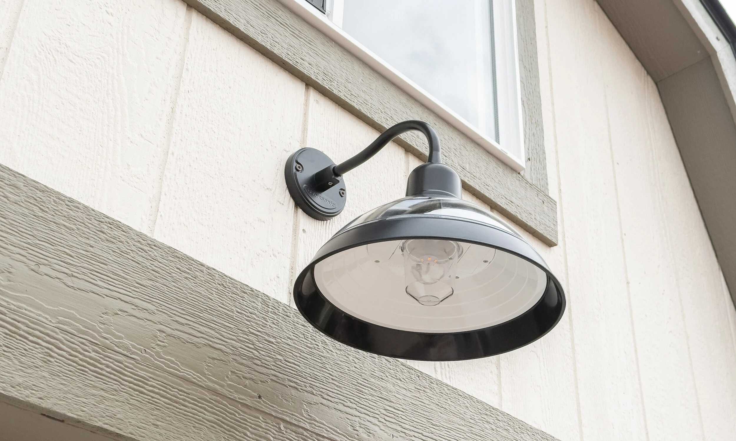 A black outdoor wall light fixture with a wide shade, mounted on a light-colored textured wall near a window.