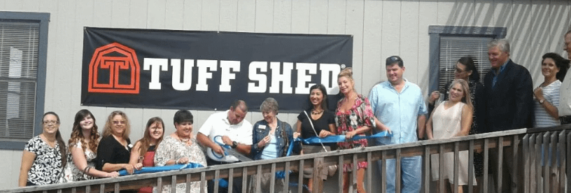 Exterior of Tuff Shed Corpus Christi location with a group of more than 12 people standing on a ramp single file slanting up to the right with one man in the middle cutting a blue ribbon with giant scissors