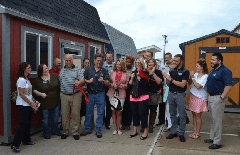 Exterior more than 15 people cutting a red ribbon with large scissors standing in a parking lot in front of a display set of Tuff Shed buildings