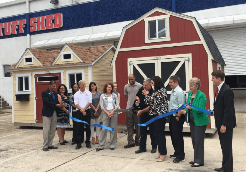 Exterior of Raleigh-Durham Tuff Shed location with 13 people cutting a blue ribbon with a large pair of scissors while standing in front of two Tuff Shed buildings