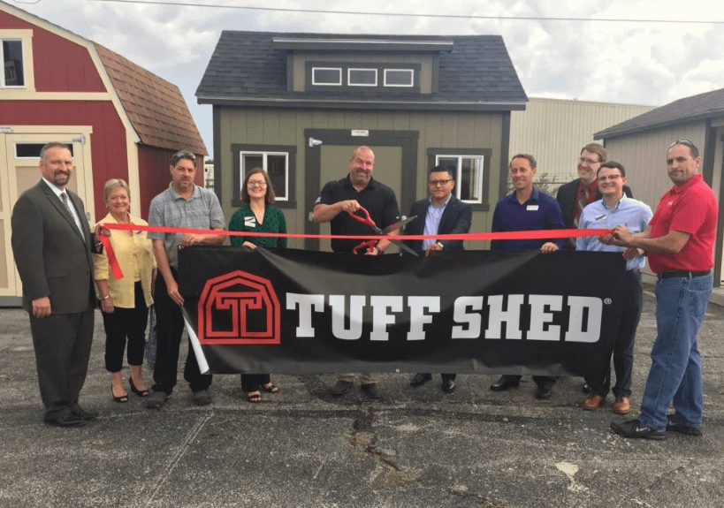 Exterior parking lot group of 10 people standing in front of a Tuff Shed, shed, holding a large Tuff Shed banner and a red ribbon which they are cutting with a large pair of scissors