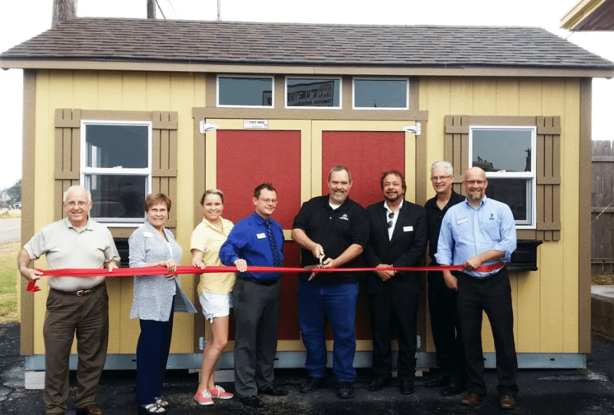 Exterior group of 8 people standing in front of a Tuff Shed, shed cutting a red ribbon for a grand opening event