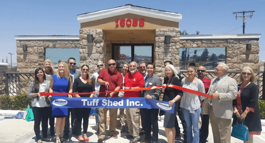 Exterior of Victorville Tuff Shed location with more than 12 people cutting a red ribbon with big scissors while holding a blue banner that says Tuff Shed Inc.
