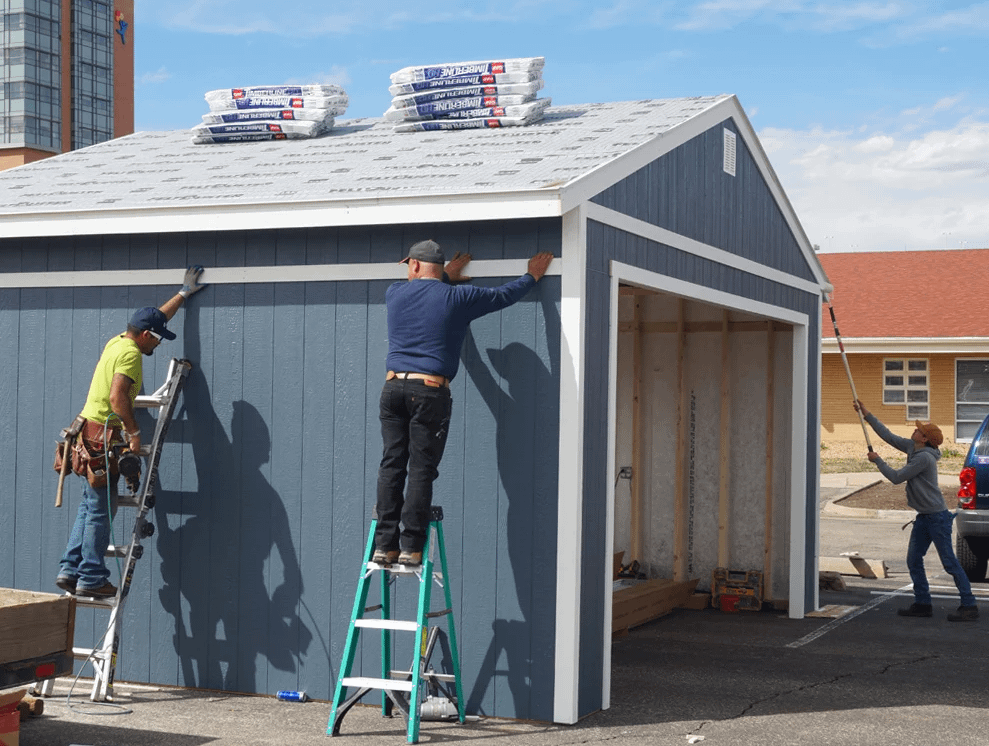 A blue Premier Ranch Garage exterior side with three installers one painting front two on ladders installing trim and tiles in packs on roof